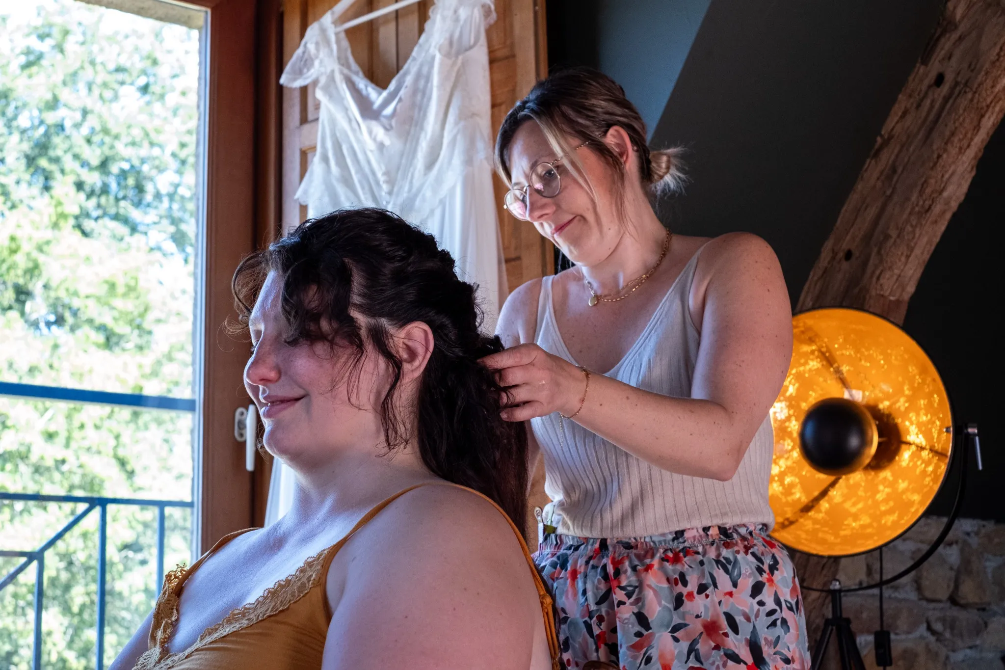 Mariée se faisant coiffer avant la cérémonie, avec sa robe en arrière-plan – Photo par Amandine Donato, photographe à Josselin, Morbihan.
