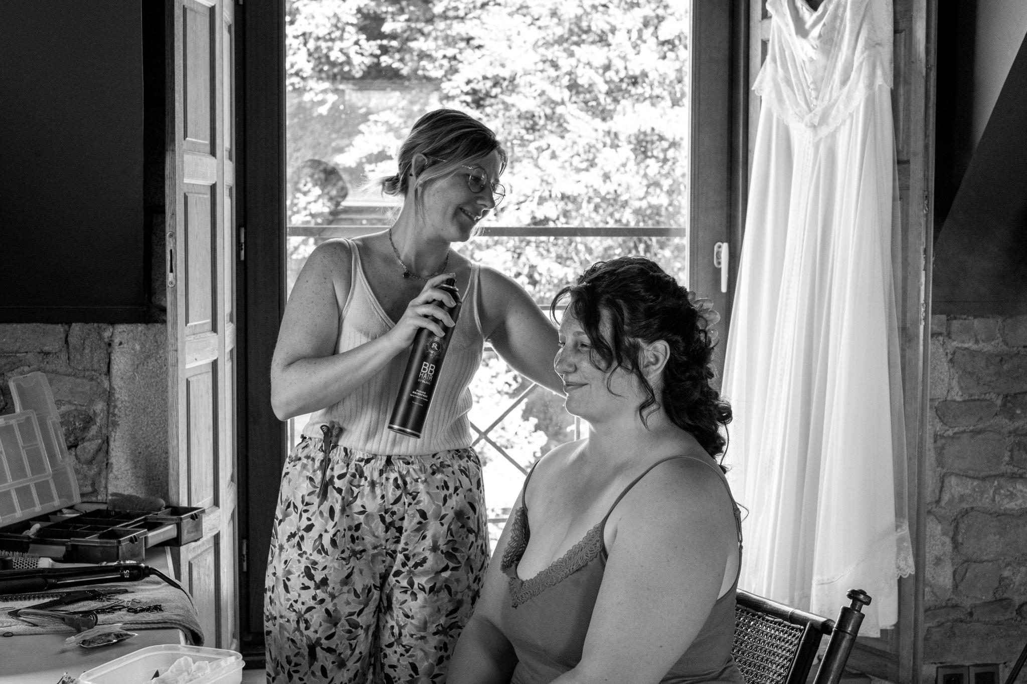 Coiffeuse ajustant la coiffure d’une mariée lors de ses préparatifs de mariage, avec sa robe en arrière-plan.