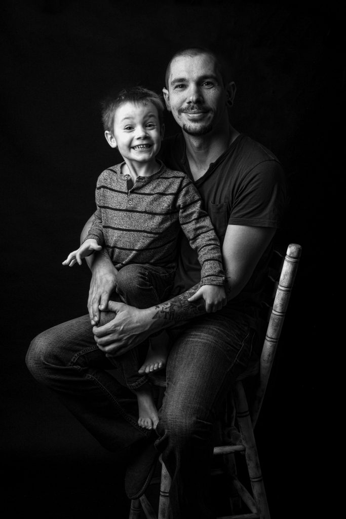 Photographie en noir et blanc d’un père tenant son jeune fils sur ses genoux pendant une séance famille en studio à Josselin, réalisée par Amandine Donato, photographe professionnelle.